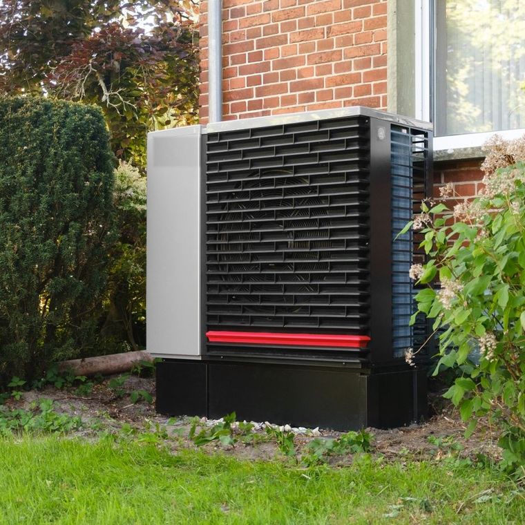 Au&szlig;eneinheit einer W&auml;rmepumpe im Garten eines Wohnhauses vor einer Backsteinfassade.