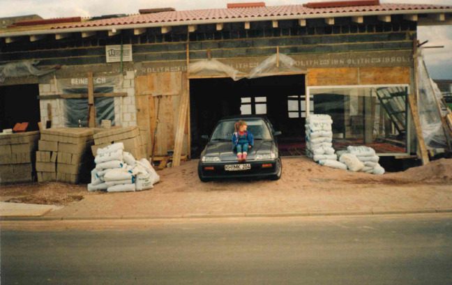 Der Rohbau des neuen Firmensitzes 1992, davor ein Auto mit einem Kind auf der Motorhaube, links und rechts gestapelte Baustoffs&auml;cke.