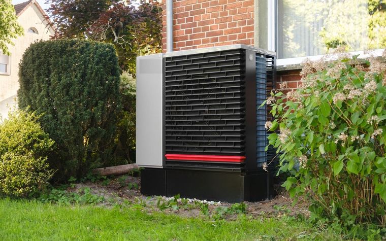 Au&szlig;eneinheit einer W&auml;rmepumpe im Garten eines Wohnhauses, dahinter eine Backsteinfassade und eine bl&uuml;hende Hecke.